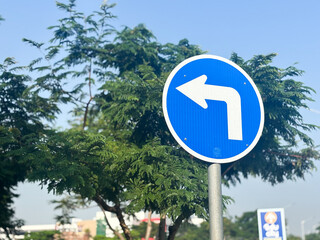 A clear, bright image of a circular blue mandatory road sign indicating a turn left ahead. The sign is mounted on a metal pole against a bright blue sky.