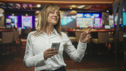 Woman holding smartphone, tapping with thumbs and smiling amid slot machines inside a casino...