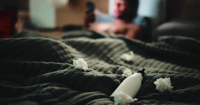 Lotion, tissues and man in bed for temptation, self pleasure and masturbation in home. Night, lubricant and person with phone for pornography, addiction and sneaky activity in bedroom of apartment