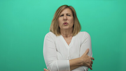 Obraz premium Woman hugs her shoulders with crossed arms and pursed lips as she shivers in studio against green backdrop; discomfort.