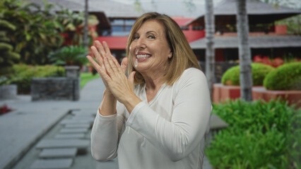 Fototapeta premium Middle aged woman smiles with closed eyes and clasps hands near chest outside resort building; gratitude.