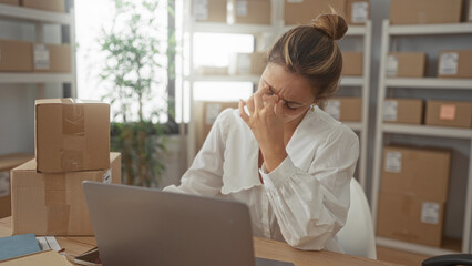 Woman rubbing eyes while holding parcel at desk in office building, removing glasses and checking laptop; stress small business struggle.