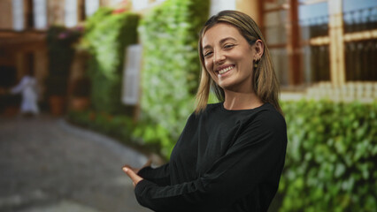 Woman smiling and gesturing with palms up toward a street building and green ivy wall, standing on...