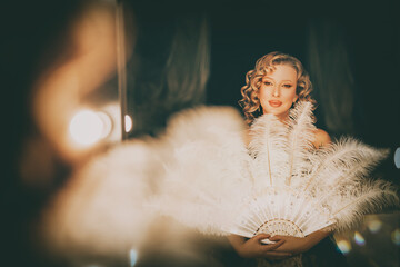 Beauty blonde hair retro lady looking into mirror hand holding fan of natural white ostrich feathers. sexy woman burlesque show actress in dressing room. Flapper girl 20s roaring twenties old style