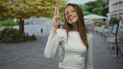 Woman points index finger forward with a smiling face on a street terrace restaurant while looking at camera; friendly greeting.