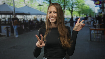 Young woman showing peace sign with both hands on a busy city street, smiling toward camera while standing near sidewalk cafes; joy positivity.