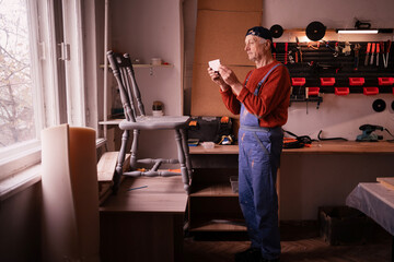 Senior craftsman in workwear restoring vintage wooden chair in workshop, photographing project with smartphone camera. Carpentry, furniture restoration, small business