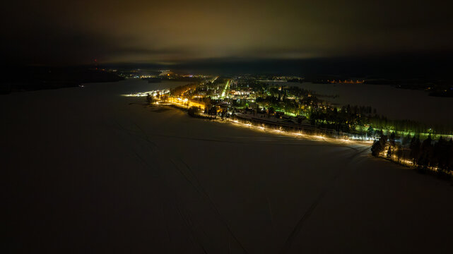 Aerial view of the dark landscape with lights illuminating the town by the still, frozen lake under the night sky, North Karelia, Finland.