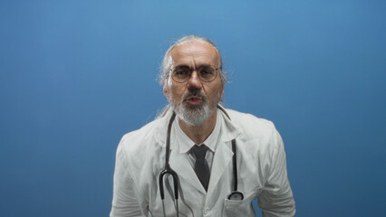 Man doctor in white coat with stethoscope pointing index fingers at camera in blue studio with long...