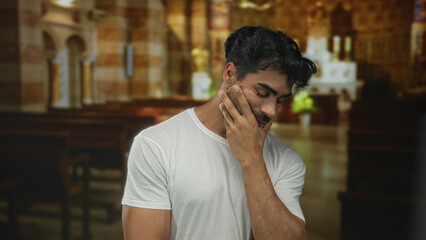 Man touching cheek and tilting head in church building with wooden pews and warm light; quiet...