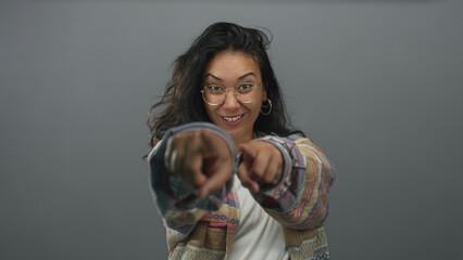 Woman points finger to camera in gray studio wearing glasses and patterned jacket, direct...