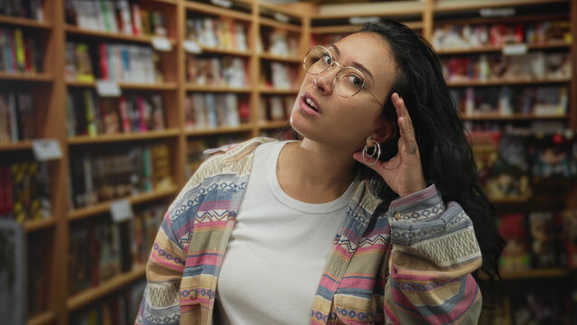 Woman cupping hand to ear and tilting head in a library, wearing glasses and patterned cardigan, surrounded by bookshelves; curiosity discovery.