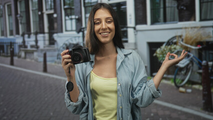 Woman holds camera near chest and makes ok sign with right hand on street lined with buildings and...