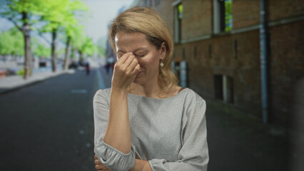 Woman folds arms and pinches nose in gray blouse on sunlit street lined with green trees and brick wall; disgust.