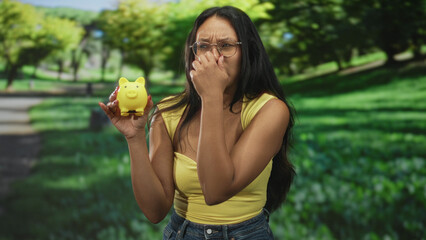 Young hispanic brunette woman wearing yellow top holding yellow piggybank pinches her nose in leafy forest; financial anxiety.