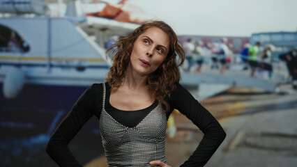 Young woman smiling and dancing happily at a lively seaside port with a yacht and blurred crowd in the background on a sunny day.