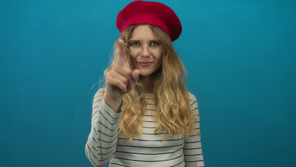 Naklejka premium Portrait of a woman whose hand is pointing at the camera in a studio as a blonde young subject wearing a beret and glasses against a blue backdrop flashes a smile.