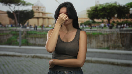 Fototapeta premium Woman pressing hand to forehead on busy cobblestone street near ancient building in bright sunlight; stress.