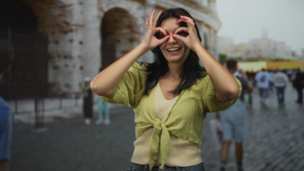 Woman makes hand binoculars gesture at coliseum building under soft daylight; curiosity exploration.