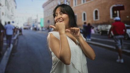 Young latina brunette woman smiling with thumb pointing sideways on a city street by brick building; playful confidence.