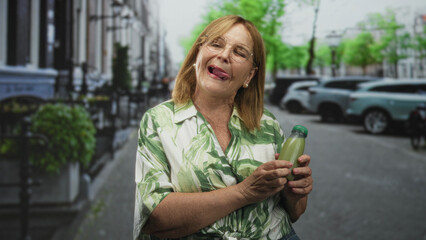 Woman holding a green juice bottle, sticks tongue out while smiling and presenting the drink on a street; playful joy.