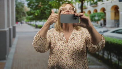 Middle age woman holding smartphone with both hands taking a photo on a street near arched building; curiosity connection.