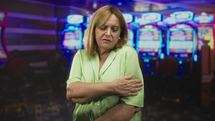 Woman clutching arm at slot machine in casino building, green blouse visible and hand on shoulder in pain; gambling distress isolation.