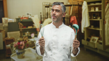 Middle age man chef with grey hair in white coat holds whisk and teal ladle in both hands and looks up in studio; quiet contemplation.