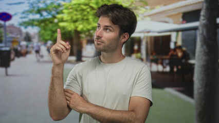 Young hispanic man points finger upward on a street terrace outside a restaurant while looking...