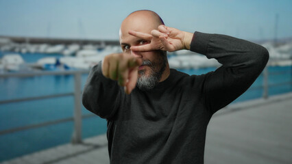 Bald bearded man making a peace gesture and pointing at the camera outdoors by a seaside port with boats and a blue sea backdrop suggesting a relaxed vacation setting.