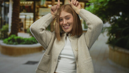 Young blonde woman wearing beige cardigan makes horns gesture with fingers on street; rock fun metal.