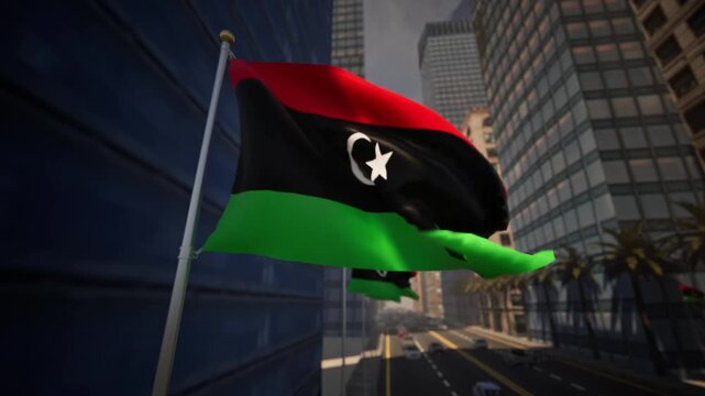 Libyan national flag waving along a modern city street under clear daylight, surrounded by buildings, palm trees and light traffic within an urban environment.
