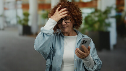 Woman holding smartphone outdoors looking worried with hand on forehead and weariness in bustling urban street environment featuring city background and day scene.