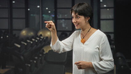 Young woman waves hand and smiles in a fitness studio, wearing an orange pendant and rings while facing mirror and rows of equipment; friendly greeting welcome.