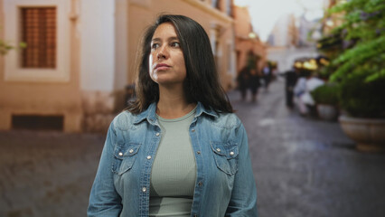 Woman in denim jacket looks left on a cobblestone street with blurred shops and planters visible,...