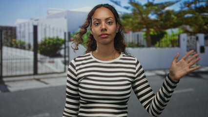 Fototapeta premium Young african american woman facing camera waving hand on a residential street outside a white building entrance; calm reflection hope.