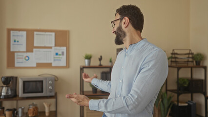 Hispanic man with beard smiling and gesturing in a modern office with charts on a corkboard,...