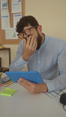 Fototapeta premium Hispanic man with a beard looks distressed while holding a blue tablet in an office setting with charts on the wall, suggesting a business situation or work stress.