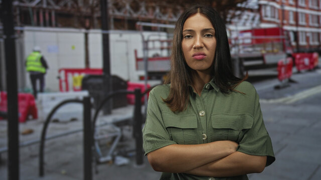 Young brunette hispanic woman with arms crossed on street at construction site beside red barriers and worker in high vis; frustration defiance.