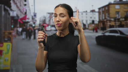 Woman holding telephone handset points finger upward on street with pursed lips and closed eyes;...