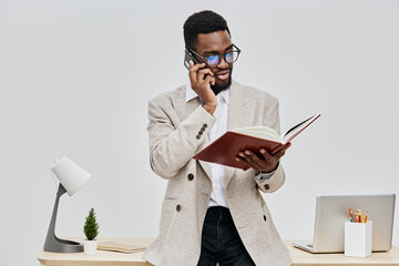 Professional Black man talking on mobile phone while reading a notebook in modern office setting, wearing beige blazer and glasses, with laptop and desk accessories, versatile workplace scene