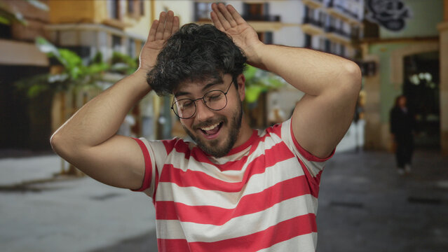 Young hispanic man in striped shirt smiling playfully with hands on head outdoors on urban street suggesting a cheerful mood