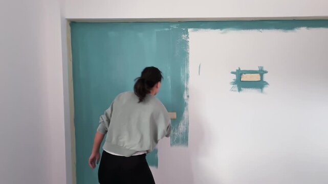 Young woman DIY painting apartment room wall with a teal color using a roller brush