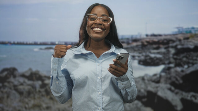 Woman holding smartphone and pumping fist while smiling in a studio with rocky seaside backdrop and eyeglasses visible; celebration success. - Powered by Adobe