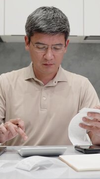 Focused adult asian man reviewing store receipt and calculating monthly expenses at home. Vertical video of budgeting, rising prices, personal finance, cost control, inflation, and money management.