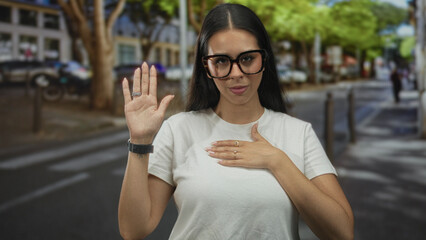 Woman with right hand raised and left hand on chest on street, wearing glasses and white tee; oath...