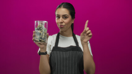 Woman brunette in striped apron holding glass jar of cash and pointing index finger in pink studio;...