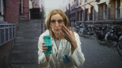 Fototapeta premium Woman doctor in lab coat holding teal mouthwash bottle, smiling mouth and pointing hand toward camera on street; cheerful promotion.