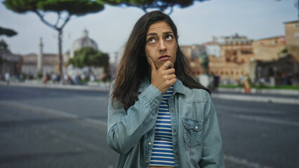 Fototapeta premium Young hispanic woman with hand on chin standing on an urban street in front of historic building; pensive pondering doubt.