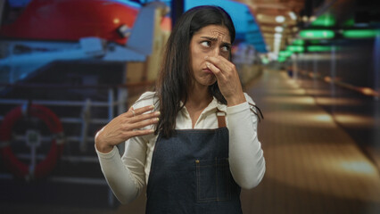 Fototapeta premium Woman pinches nose and covers mouth with hand wearing denim apron on studio boat deck set with visible lifebuoy and railing; disgust smell complaint.